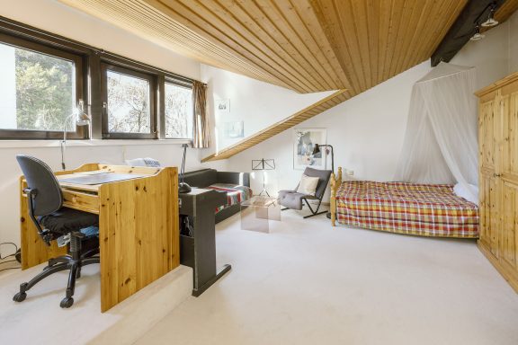 Ein helles Zimmer mit einem Schreibtisch, Stuhl, Bett und Holzregalen.