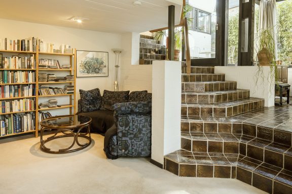 Gemütlicher Wohnraum mit Bücherregal, schwarzem Sofa und Treppe aus Fliesen.