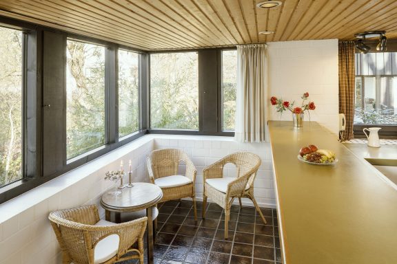 Helle Wohnküche mit Holzdecke, zwei Stühlen und einem Tisch, umgeben von Fenstern.